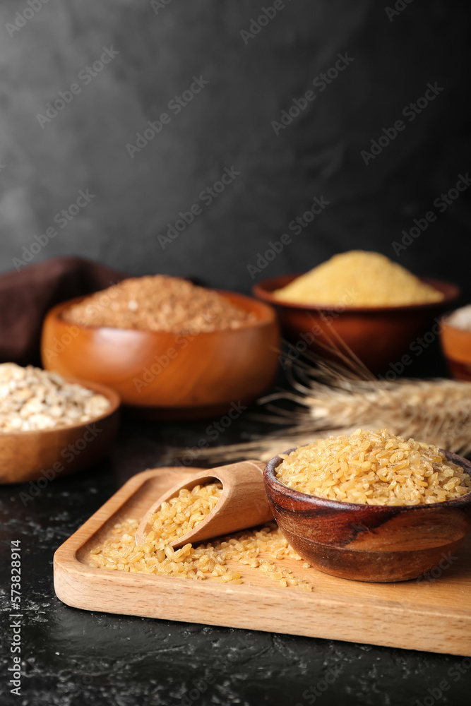 Wooden board with bowl and scoop of bulgur on dark background