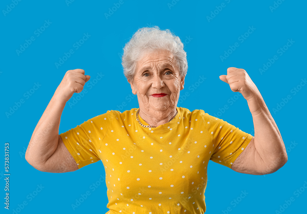 Senior woman in yellow t-shirt showing muscles on blue background