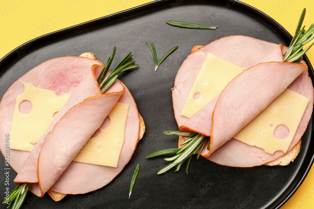 Board with slices of tasty ham, cheese, rosemary and bread on yellow background