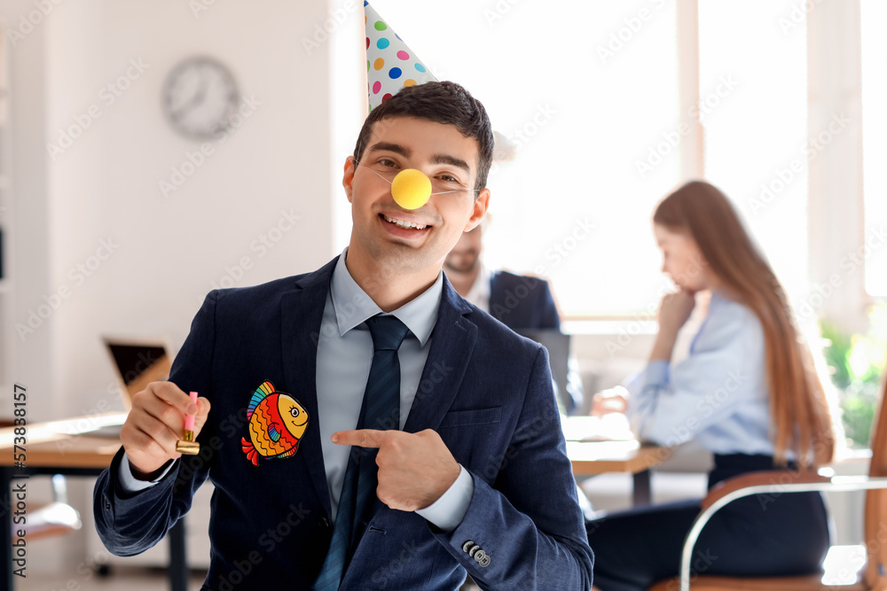 Young man with party hat and clown nose pointing at paper fish in office. April Fools Day celebrati