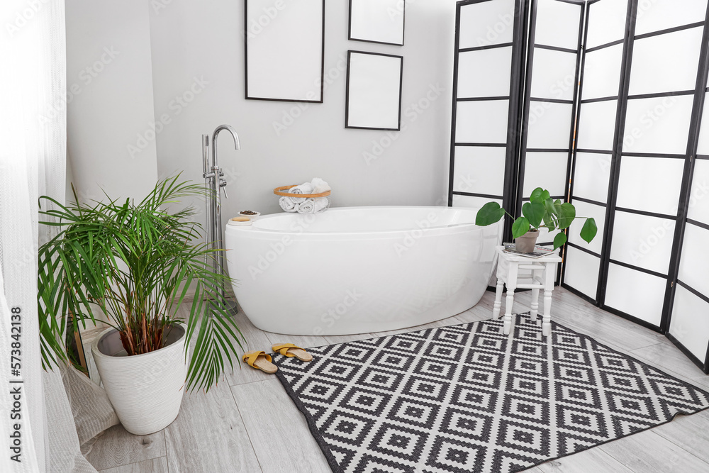 Interior of light bathroom with bathtub, blank frames and houseplants