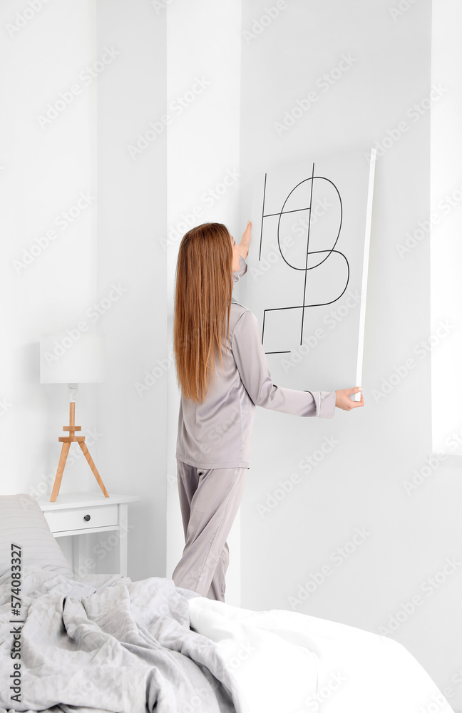 Young woman hanging poster on light wall in bedroom