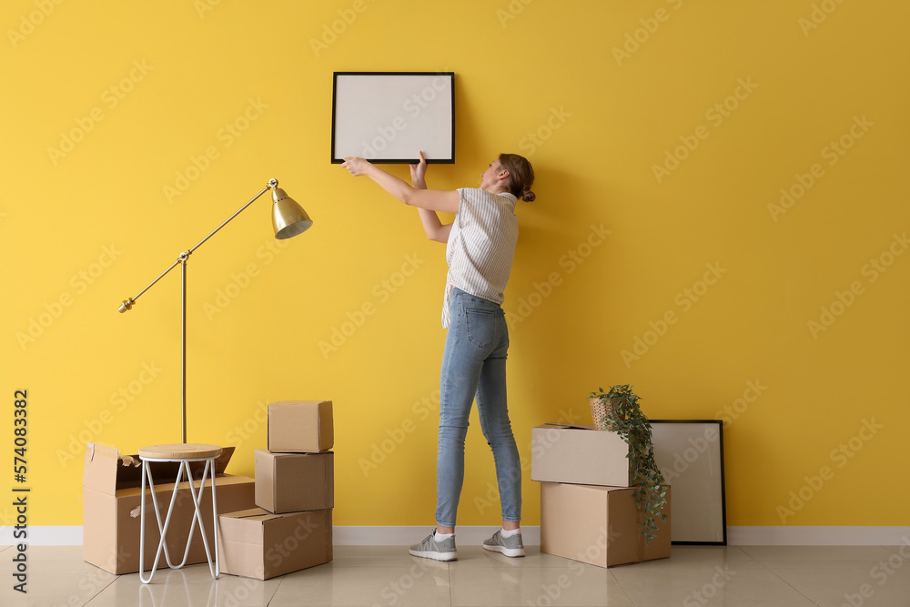 Young woman hanging blank frame on yellow wall at home