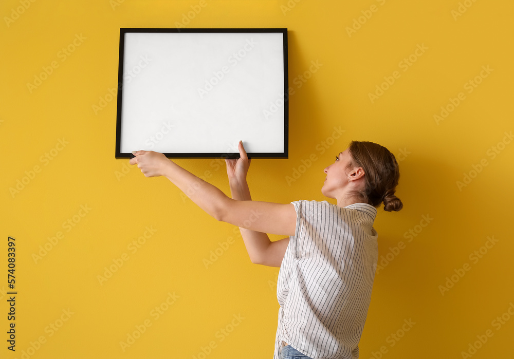 Young woman hanging blank frame on yellow wall at home