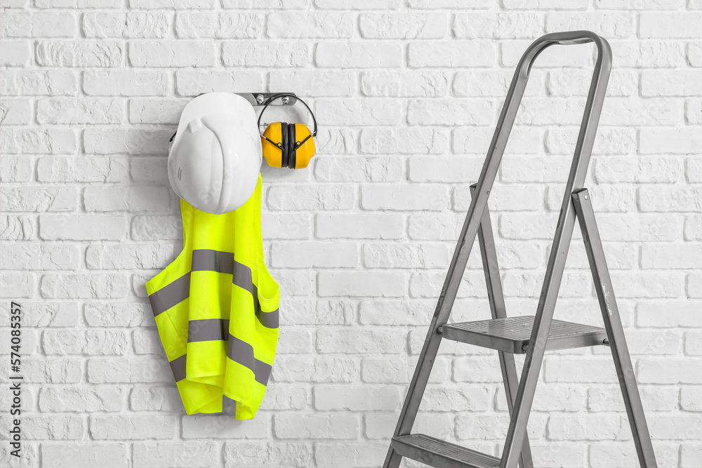 Stepladder and reflective vest with hearing protectors on white brick wall