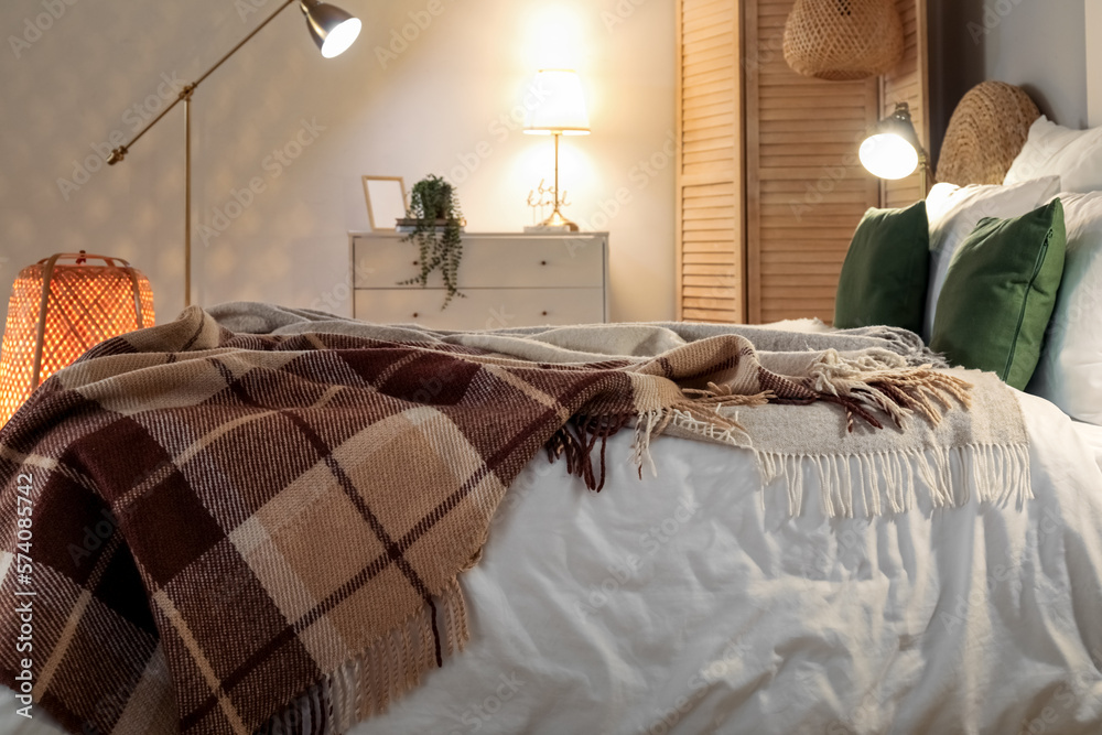 Interior of bedroom with brown checkered blankets on bed and glowing lamps late in evening