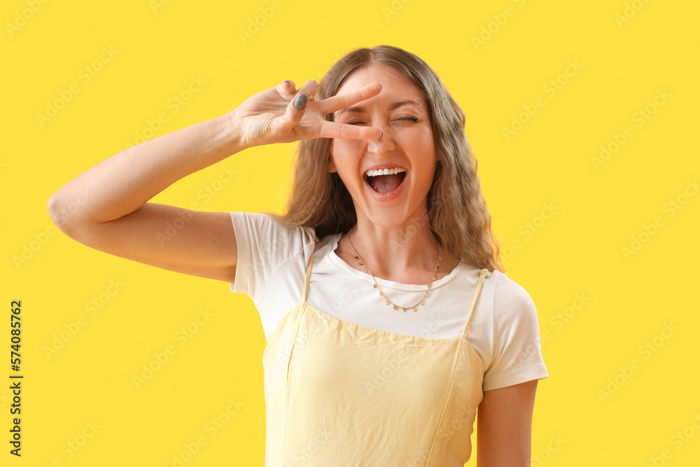 Happy young woman showing victory gesture on yellow background