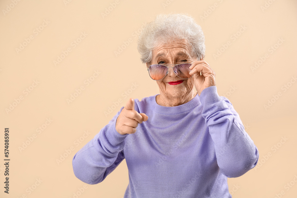 Senior woman in sunglasses pointing at viewer on beige background