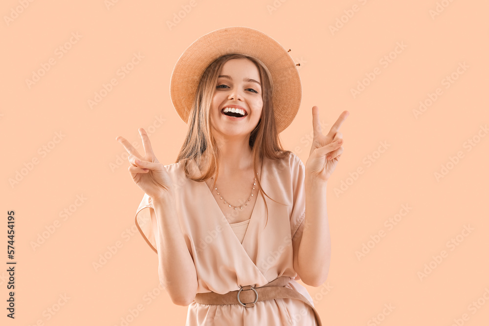 Young woman in hat showing victory gesture on beige background