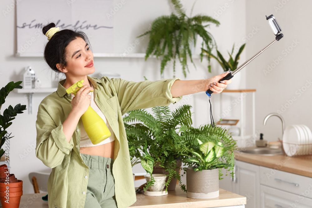 Young woman with spray bottle and green houseplants taking selfie in kitchen