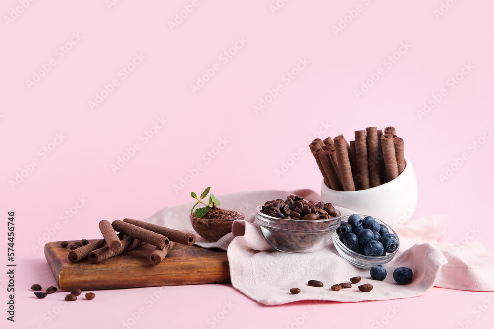 Board with delicious chocolate wafer rolls, coffee beans and blueberries on pink background