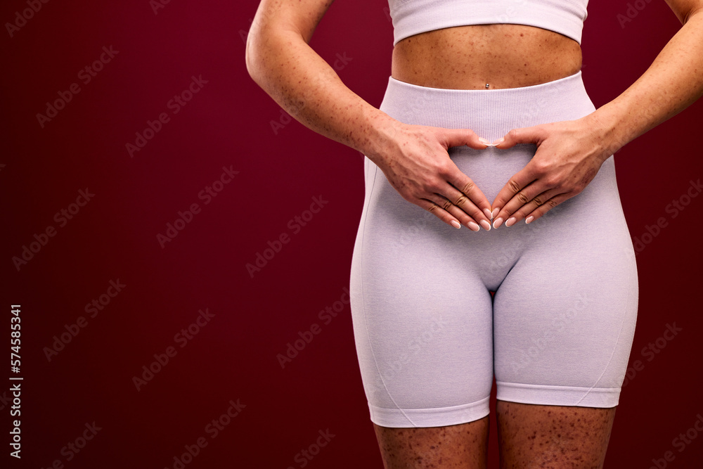 A woman keeps her hands in a heart shape on her stomach, has spotted skin, feeling confident.