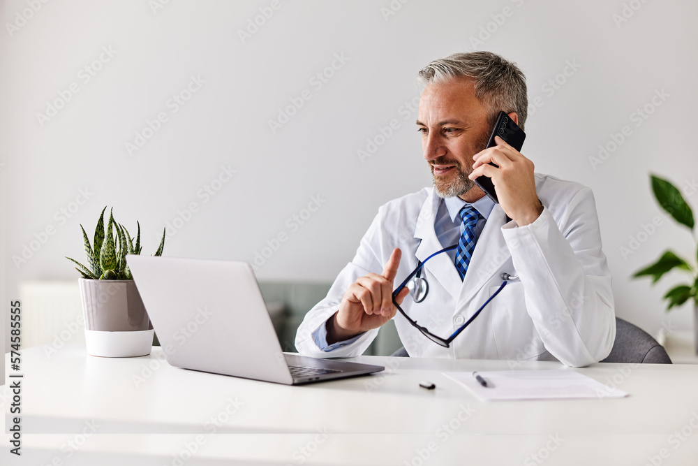 A serious male doctor is talking over the phone and working on the laptop at the office.