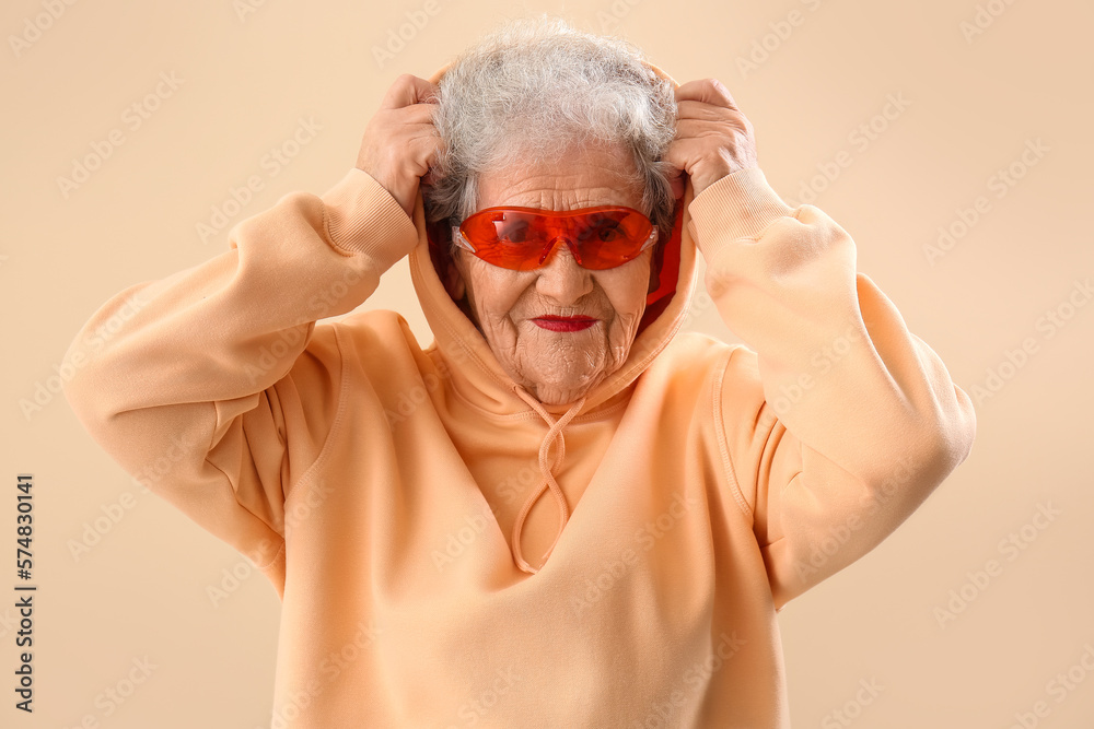 Senior woman in sunglasses and hoodie on beige background