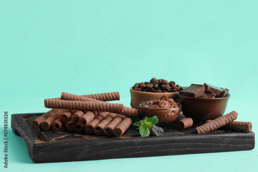 Black wooden board with delicious wafer rolls, chocolate and coffee beans on turquoise background