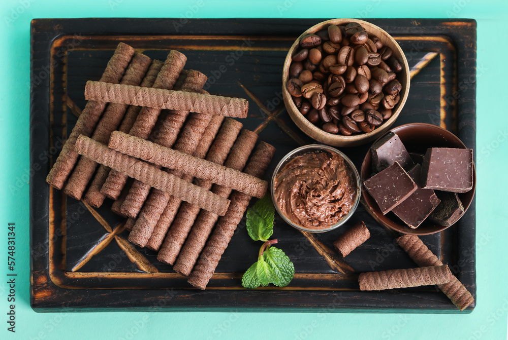 Black wooden board with delicious wafer rolls, chocolate and coffee beans on turquoise background