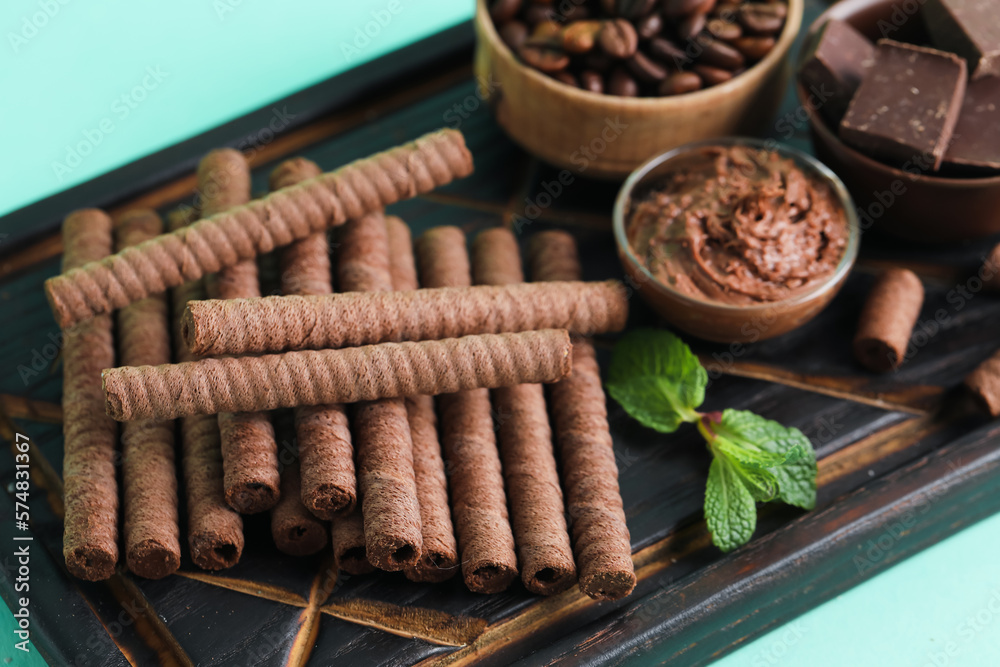 Black wooden board with delicious wafer rolls, chocolate and coffee beans on turquoise background