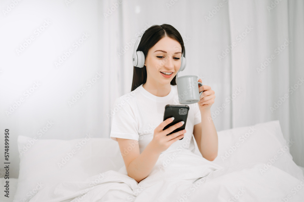 Beautiful young woman on a white bed with coffee cub and using smart phone at home. lifestyle concep