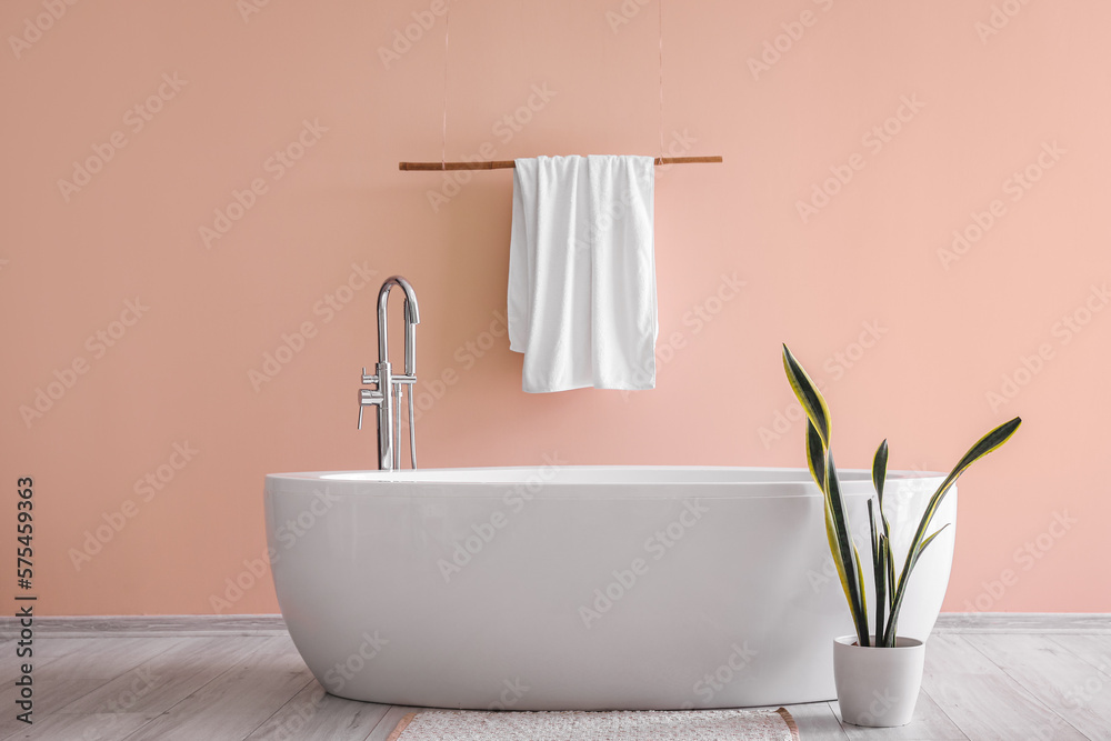 Modern bathtub with towel and houseplant near pink wall