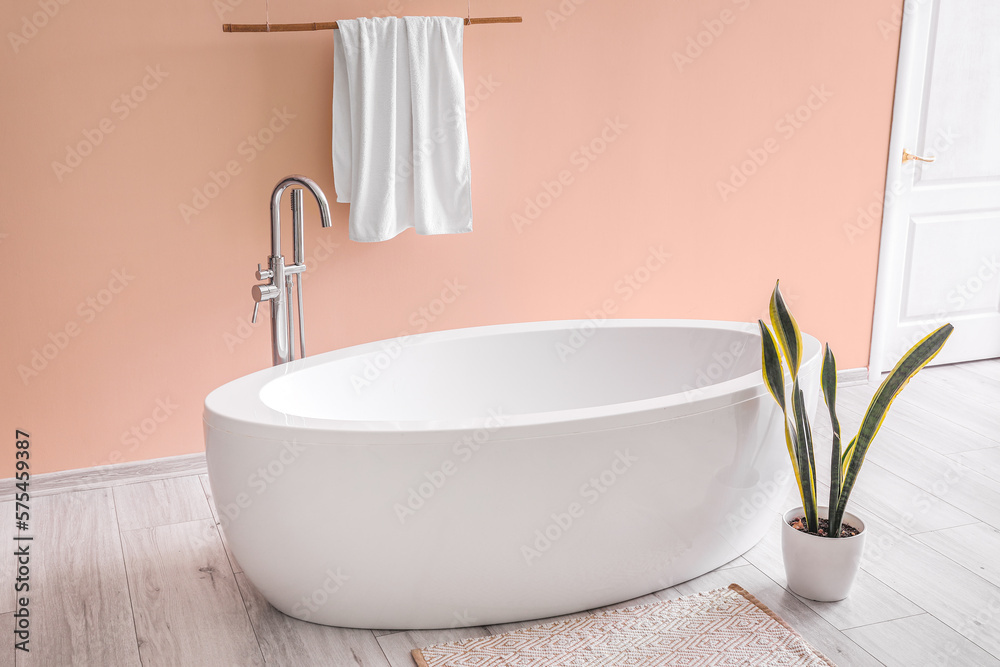 Modern bathtub with towel and houseplant near pink wall