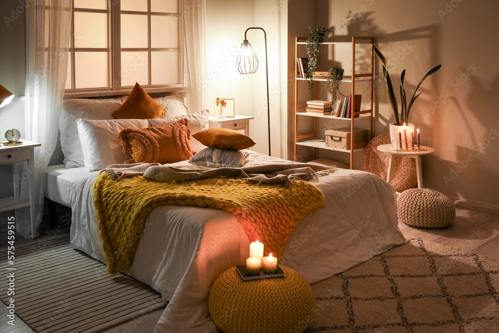 Interior of bedroom with cozy blankets on bed, burning candles and glowing lamp late in evening