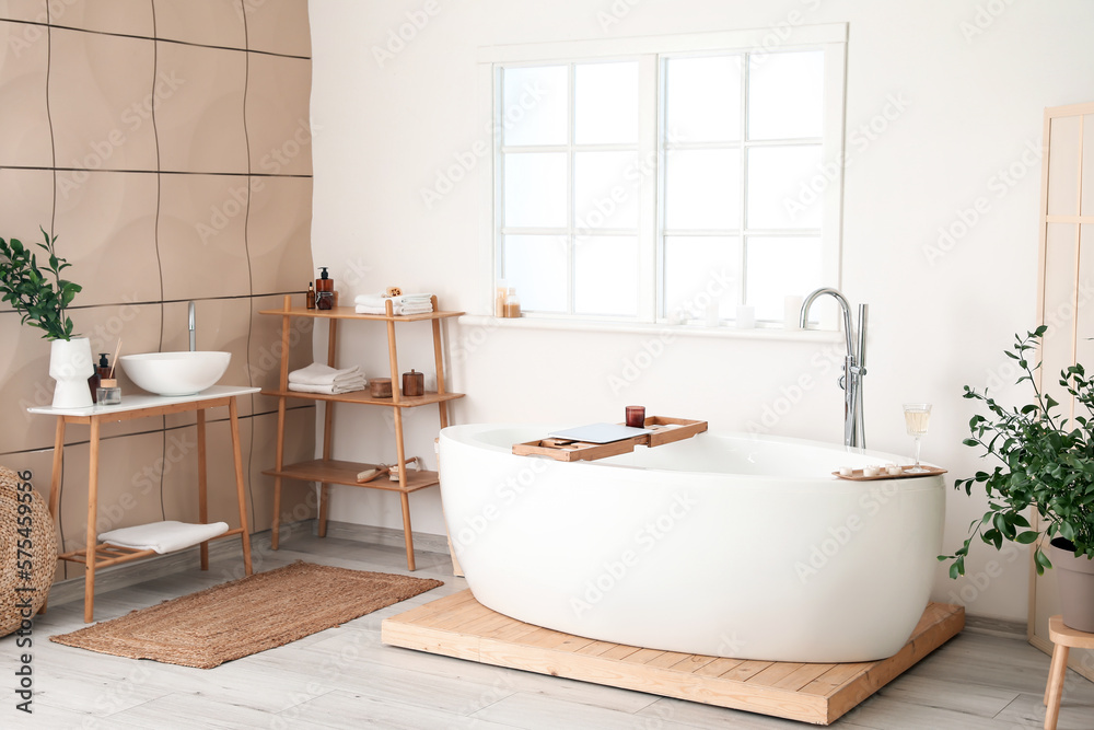 Interior of bathroom with modern bathtub, laptop, champagne and burning candles on board