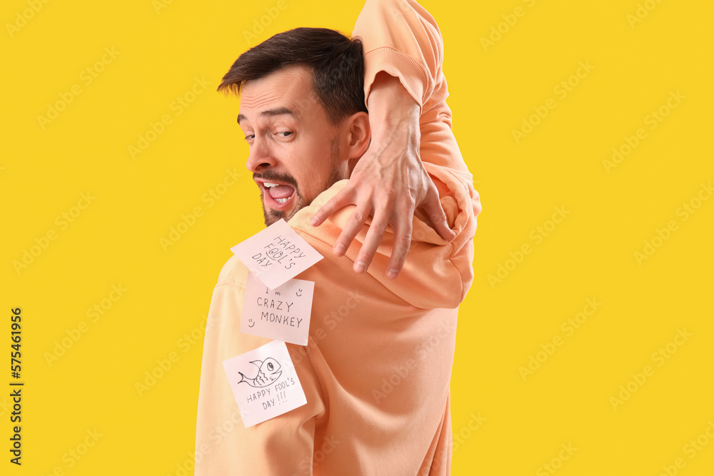 Angry young man with sticky papers on his back against yellow background. April Fools Day celebrati