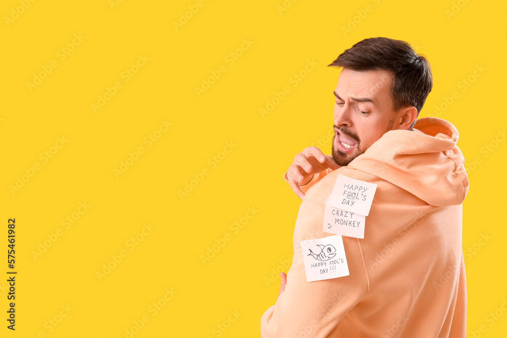 Angry young man with sticky papers on his back against yellow background. April Fools Day celebrati