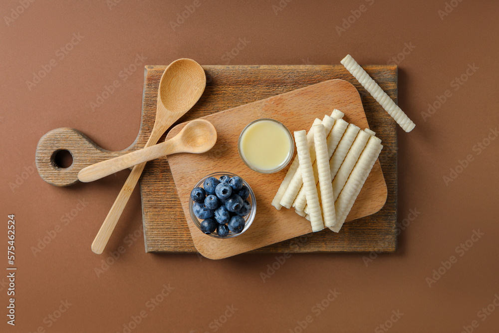 Boards with delicious wafer rolls, condensed milk and blueberries on brown background