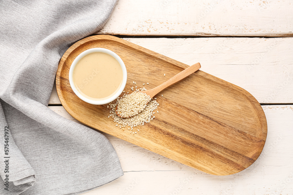 Board with bowl of tasty tahini and sesame seeds on light wooden background