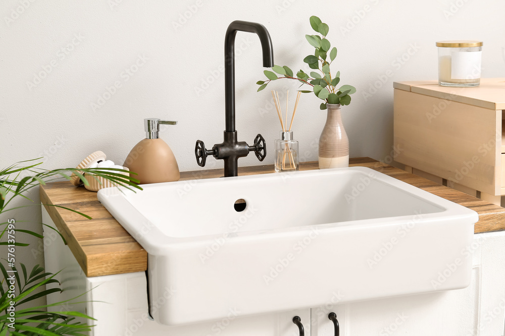 Ceramic sink with bath accessories, reed diffuser and eucalyptus in vase near light wall