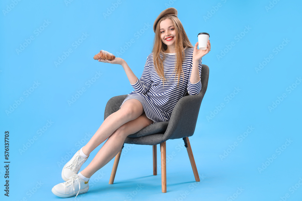 Young woman with croissant and cup of coffee sitting in armchair on blue background