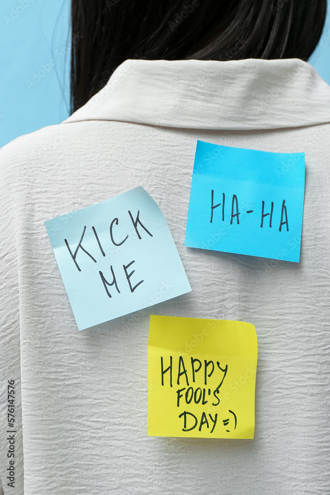 Sticky papers on womans back against blue background, closeup. April Fools Day celebration