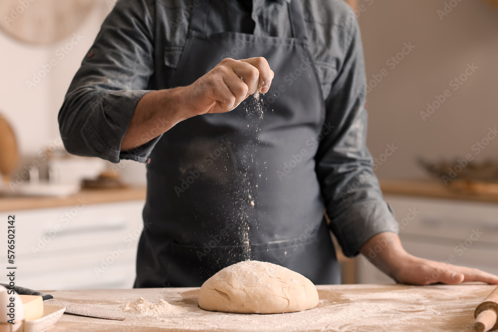 Male baker sprinkling dough with flour at table in kitchen, closeup