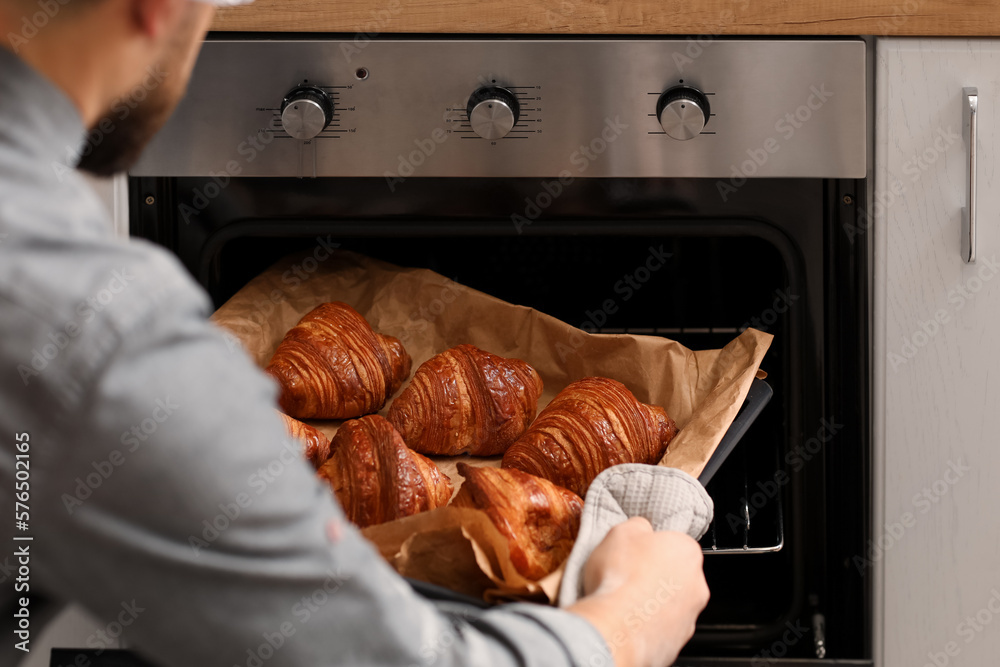 Male baker taking tray with croissants from oven in kitchen, closeup