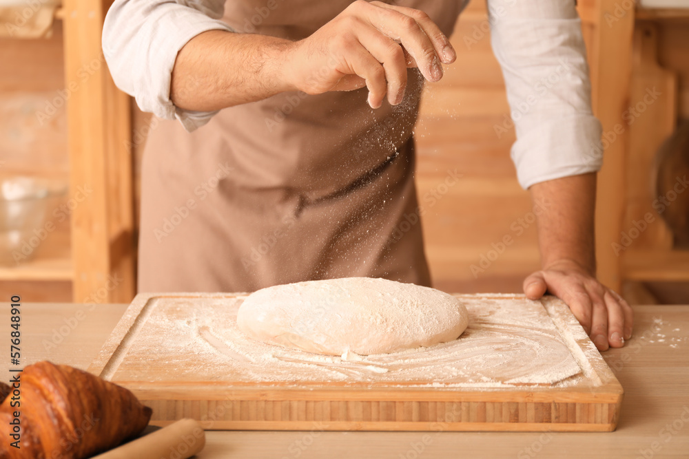 Male baker sprinkling dough with flour at table in kitchen, closeup