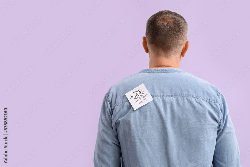 Sticky paper with text HAPPY FOOLS DAY on back of mature man against lilac background