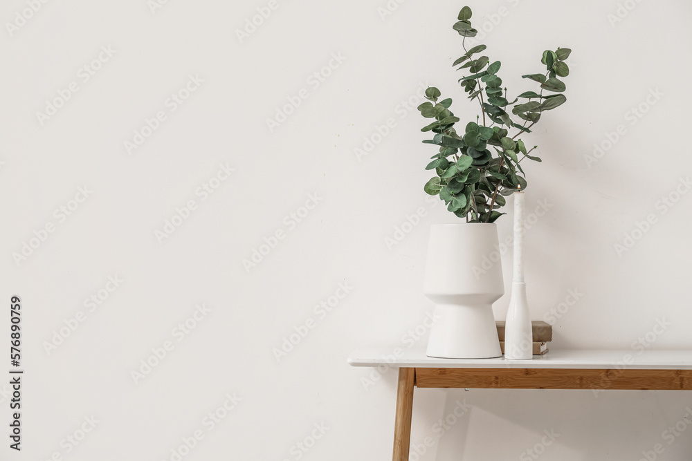 Vase with eucalyptus branches, burning candle and books on table near light wall