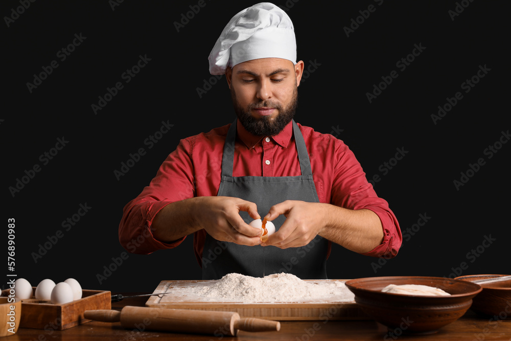 Male baker breaking egg for dough at table on dark background