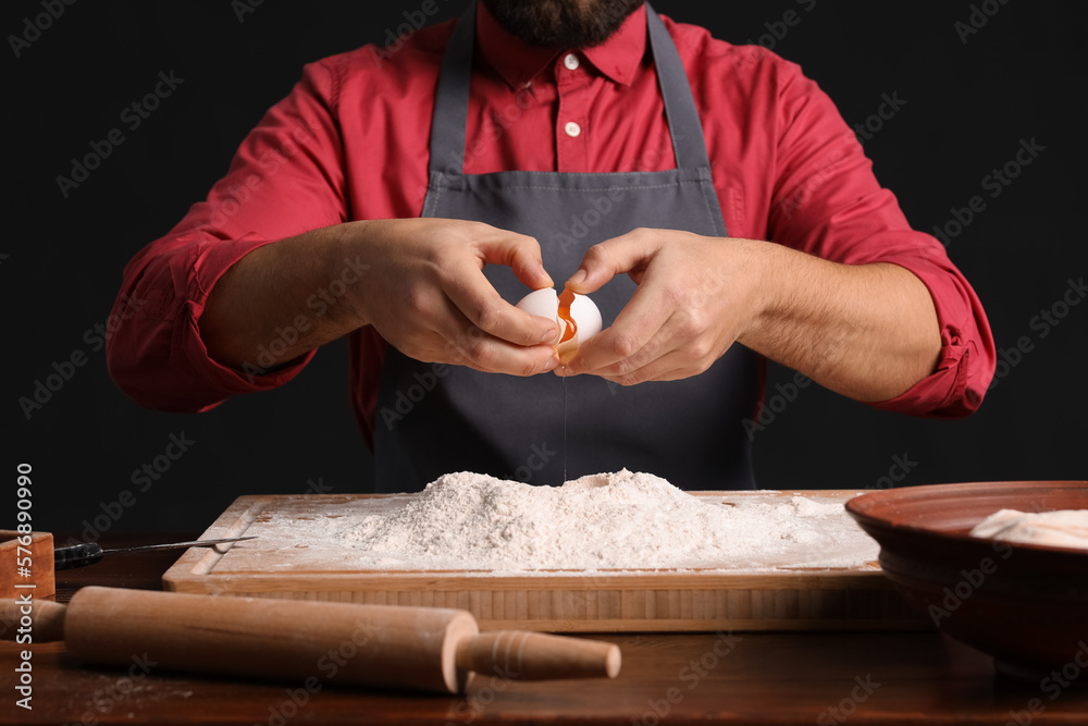 Male baker breaking egg for dough at table on dark background, closeup