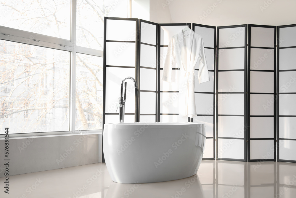 Interior of room with modern bathtub, folding screen and bathrobe near big window