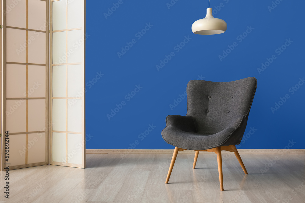 Stylish grey armchair, lamp and folding screen near blue wall