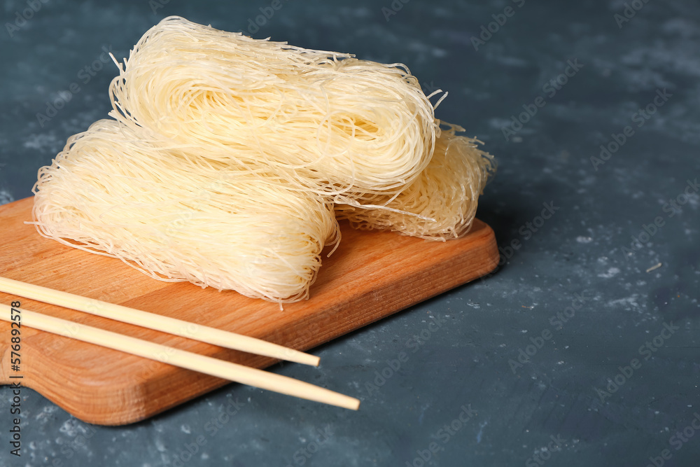 Wooden board with raw rice noodles and chopsticks on dark background