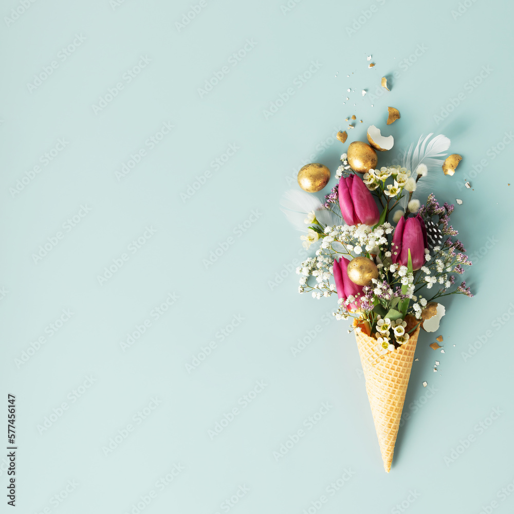 Ice cream cone with beautiful flowers and golden easter eggs on blue background flat lay