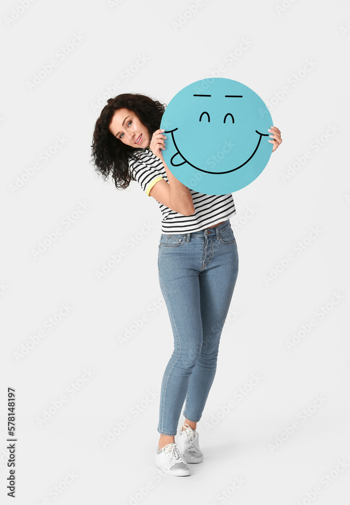 Happy young woman holding paper with smiling emoticon on light background