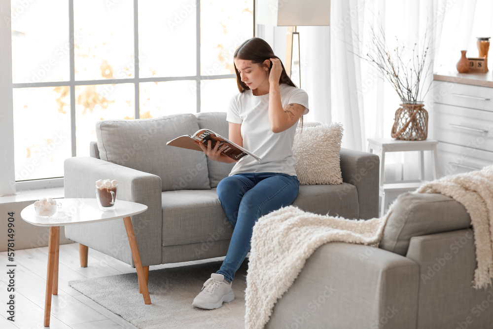 Young woman reading magazine on grey sofa in kitchen