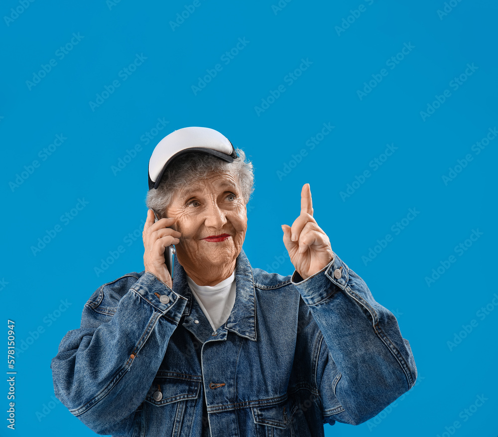 Senior woman in cap with mobile phone pointing at something on blue background