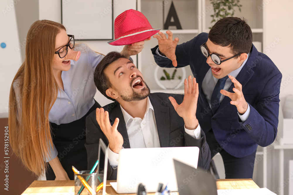 Colleagues with funny disguise putting hat on their boss in office. April Fools Day celebration