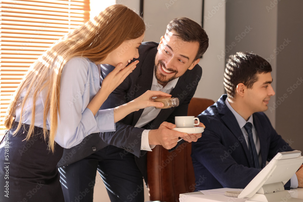 Young man and woman sprinkling pepper into their colleagues cup of coffee in office. April Fools D