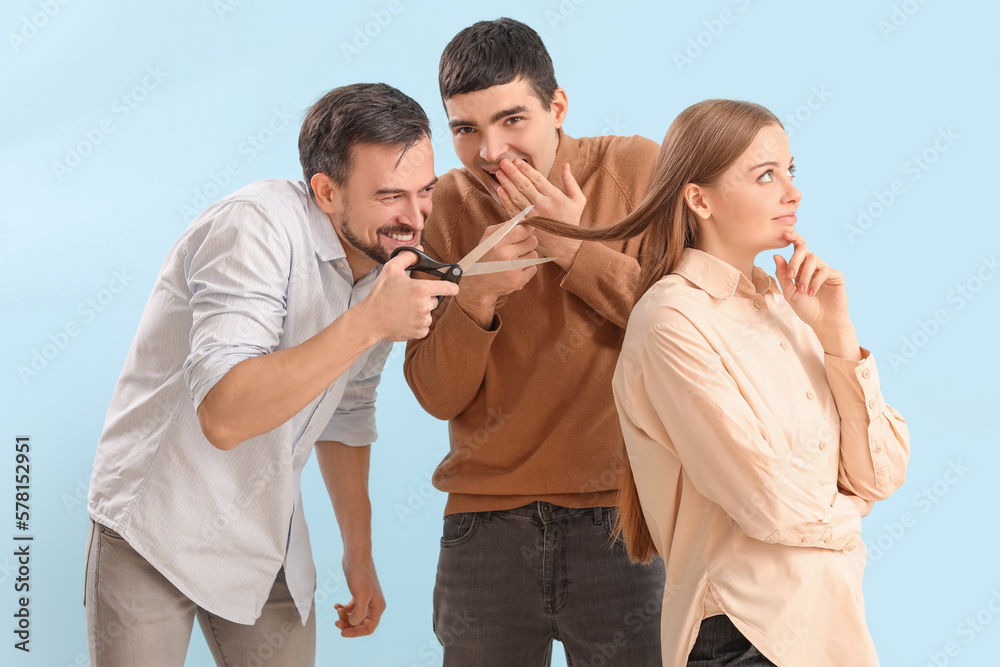 Young men with scissors cutting hair of their colleague on blue background. April Fools Day prank